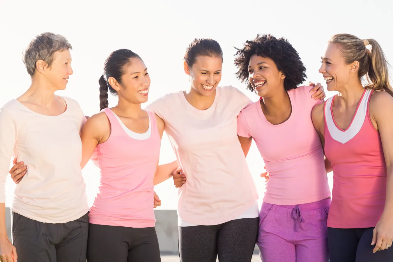 Five women in pink shirts standing together, smiling and posing for the camera in a friendly group setting.